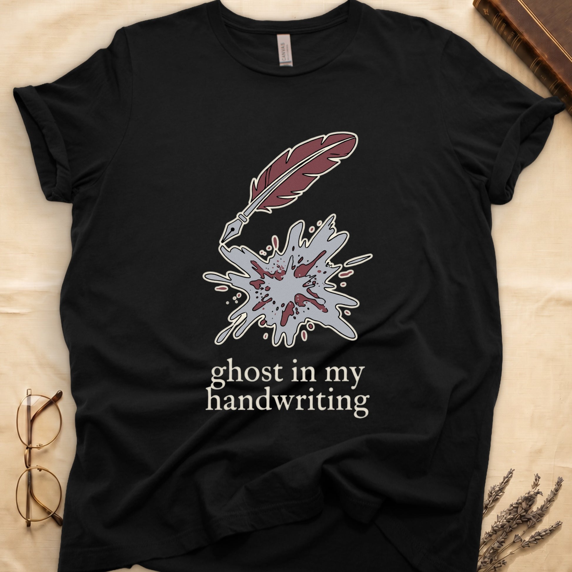 Black tee with burgundy quill pen and grey ink splatter, "ghost in my handwriting" cream serif below.