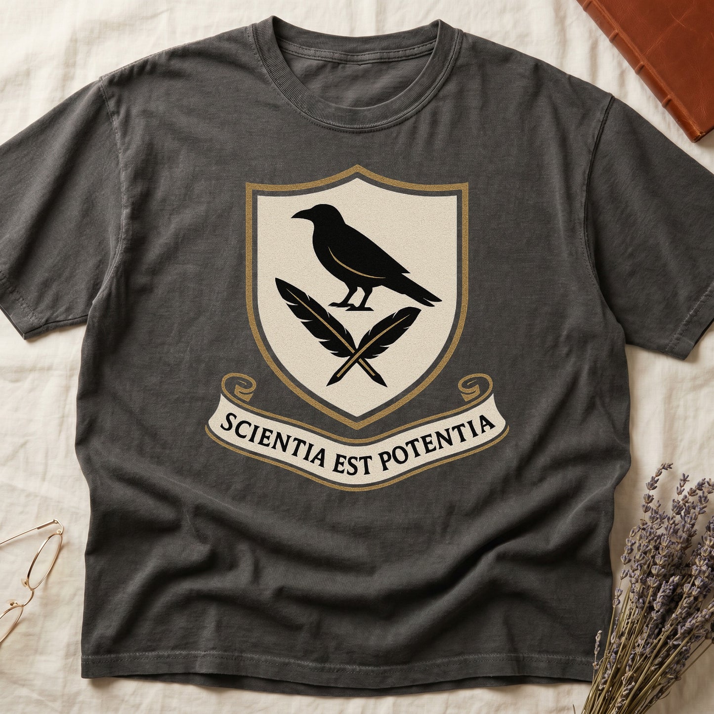 Pepper tee with cream-and-gold heraldic shield, black raven over crossed quills, scroll banner "SCIENTIA EST POTENTIA" below.