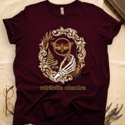 Maroon tee with skeletal hand, magnifying glass, death's-head moth, ferns on green, "mīrābilia obscūra" in Latin italic.