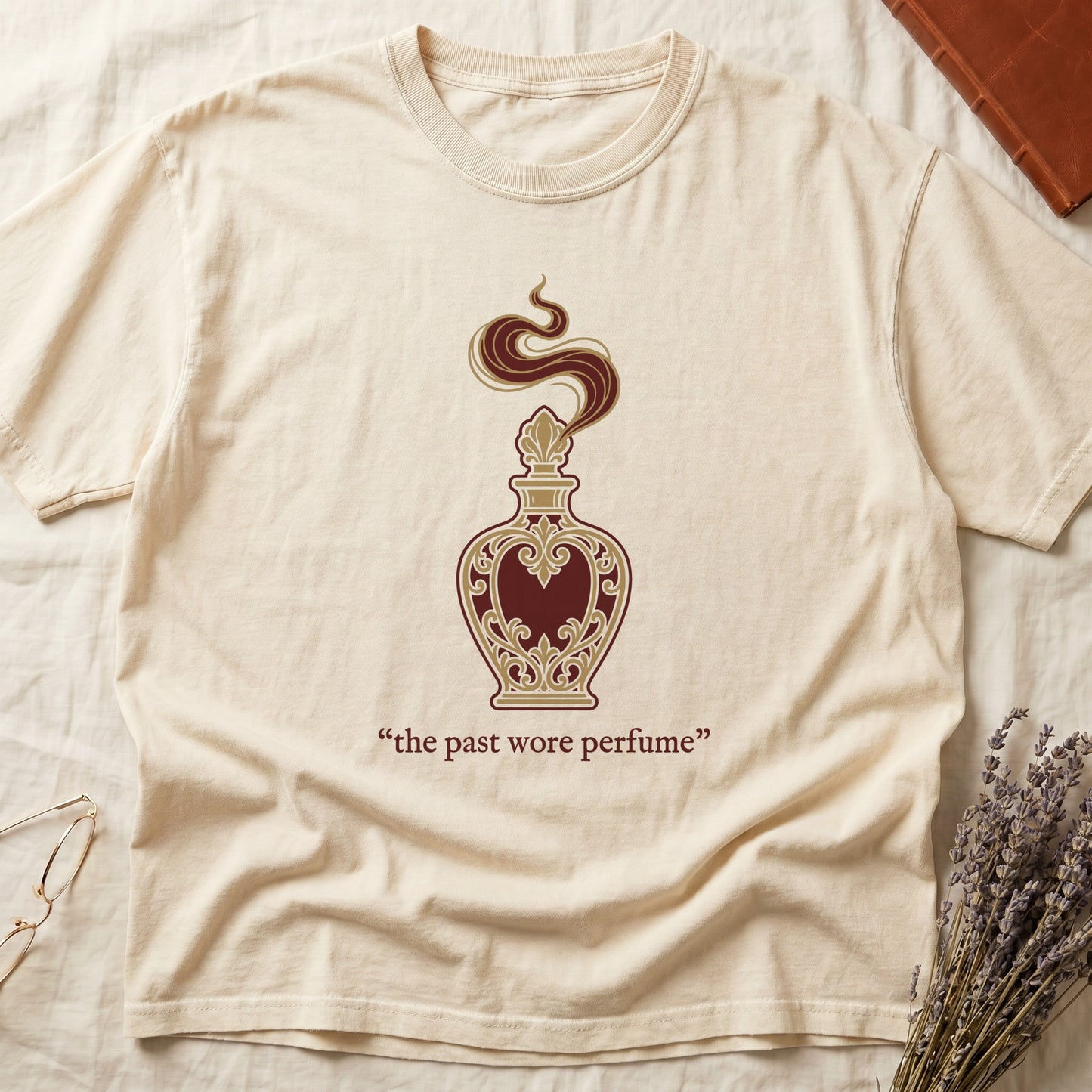 Ivory tee with ornate blue and gold perfume bottle, rising scent swirl, "the past wore perfume" cream serif below.