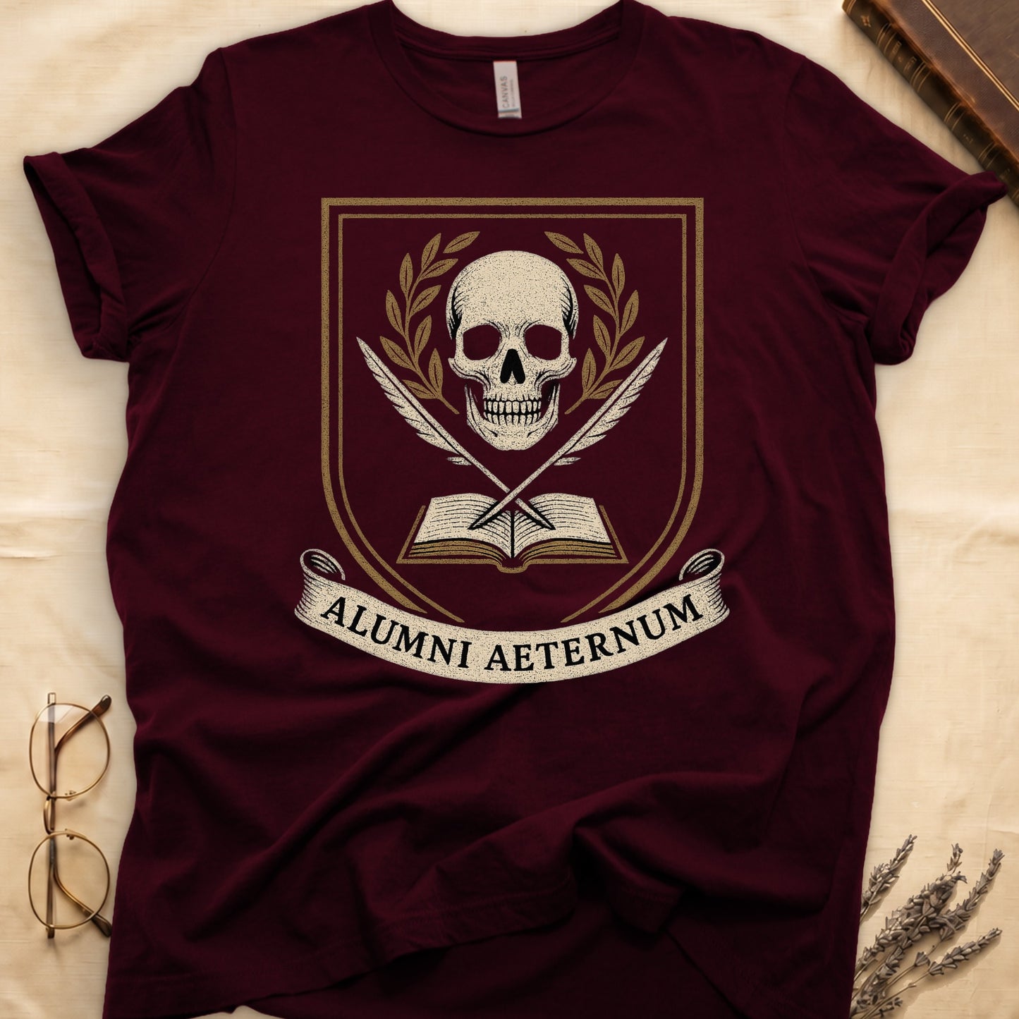 Maroon tee with heraldic shield, skull and gold laurels, crossed quills, open book, scroll "ALUMNI AETERNUM" below.