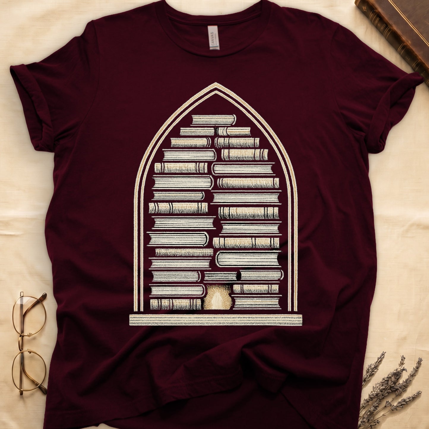 Maroon tee with gothic arch frame, columns of leather-bound books inside, sunburst medallion at base, cream engraved linework.