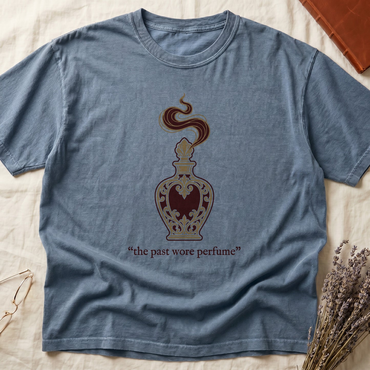 Blue Jean tee with ornate blue and gold perfume bottle, rising scent swirl, "the past wore perfume" cream serif below.