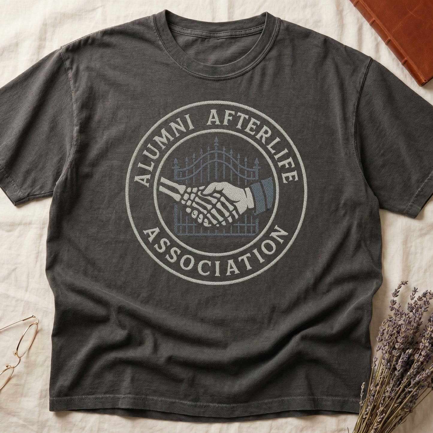 Green tee with embroidered patch, skeletal handshake at cemetery gate, "ALUMNI AFTERLIFE" top, "ASSOCIATION" bottom.