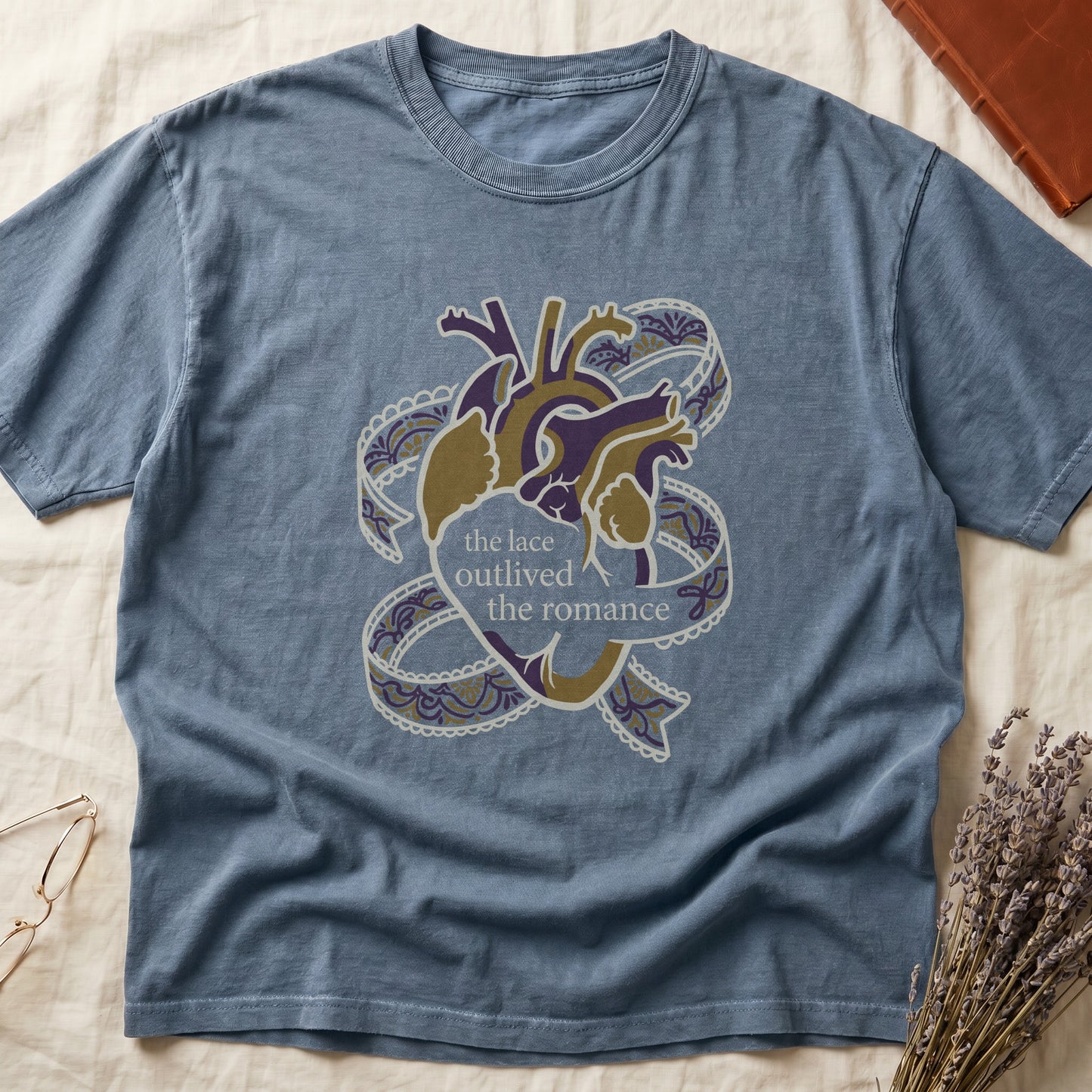 Blue Jean tee with anatomical heart in purple and gold wrapped in lace ribbons, "the lace outlived the romance" in serif.