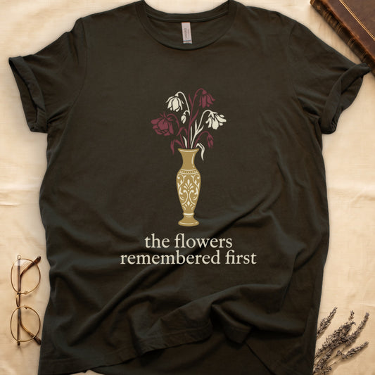Military Green tee with gold ornate vase, wilting cream lilies and dark red roses, "the flowers remembered first" in cream serif.