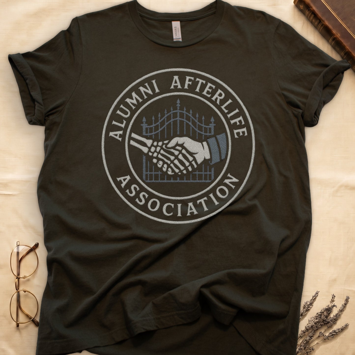Maroon tee with embroidered patch, skeletal handshake at cemetery gate, "ALUMNI AFTERLIFE" top, "ASSOCIATION" bottom.