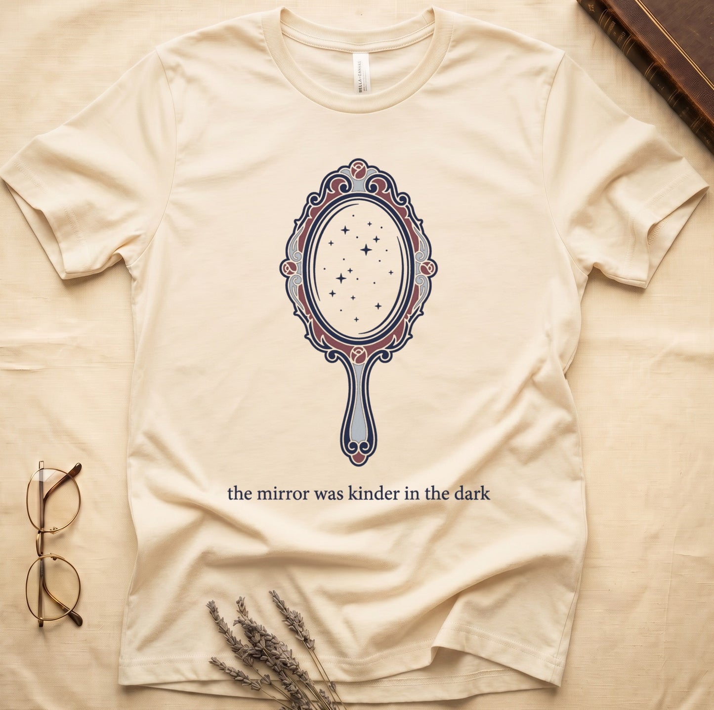 Natural tee with ornate hand mirror, dark red roses, starfield reflection, "the mirror was kinder in the dark" serif below.