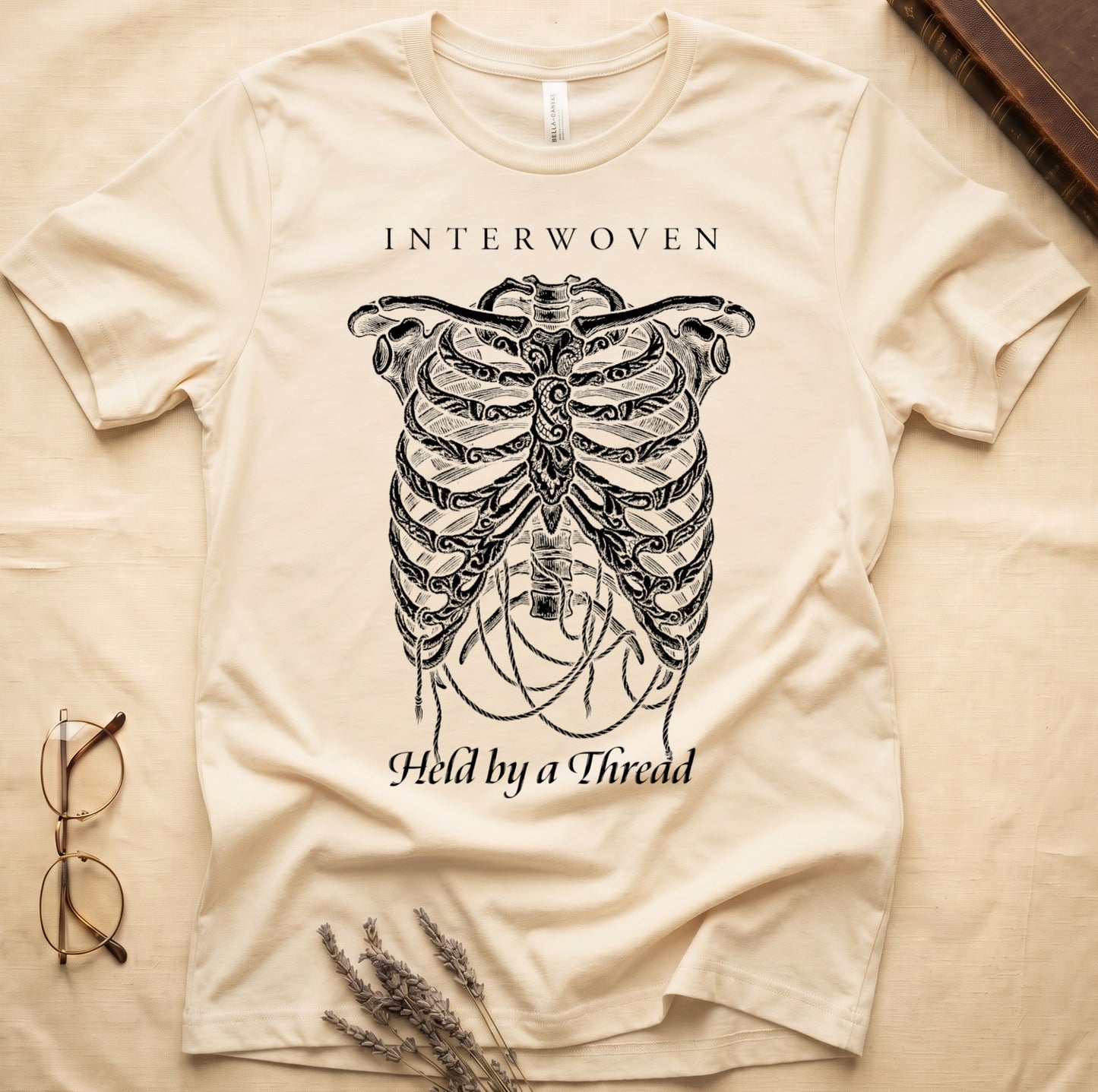 Natural tee with cream skeletal ribcage suspended by trailing threads, "Held by a Thread" in serif on plate below.