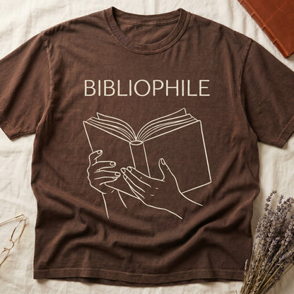 Espresso tee with two hands cradling an open book in cream and brown linework, "BIBLIOPHILE" in serif capitals above.