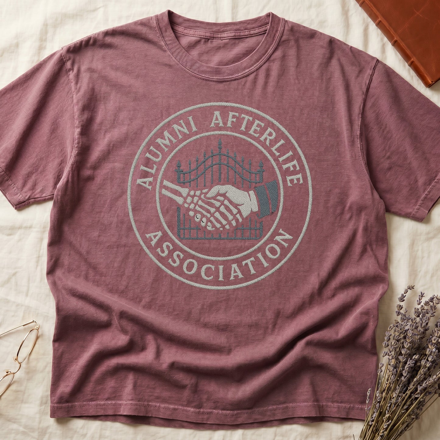 Blue Jean tee with embroidered patch, skeletal handshake at cemetery gate, "ALUMNI AFTERLIFE" top, "ASSOCIATION" bottom.