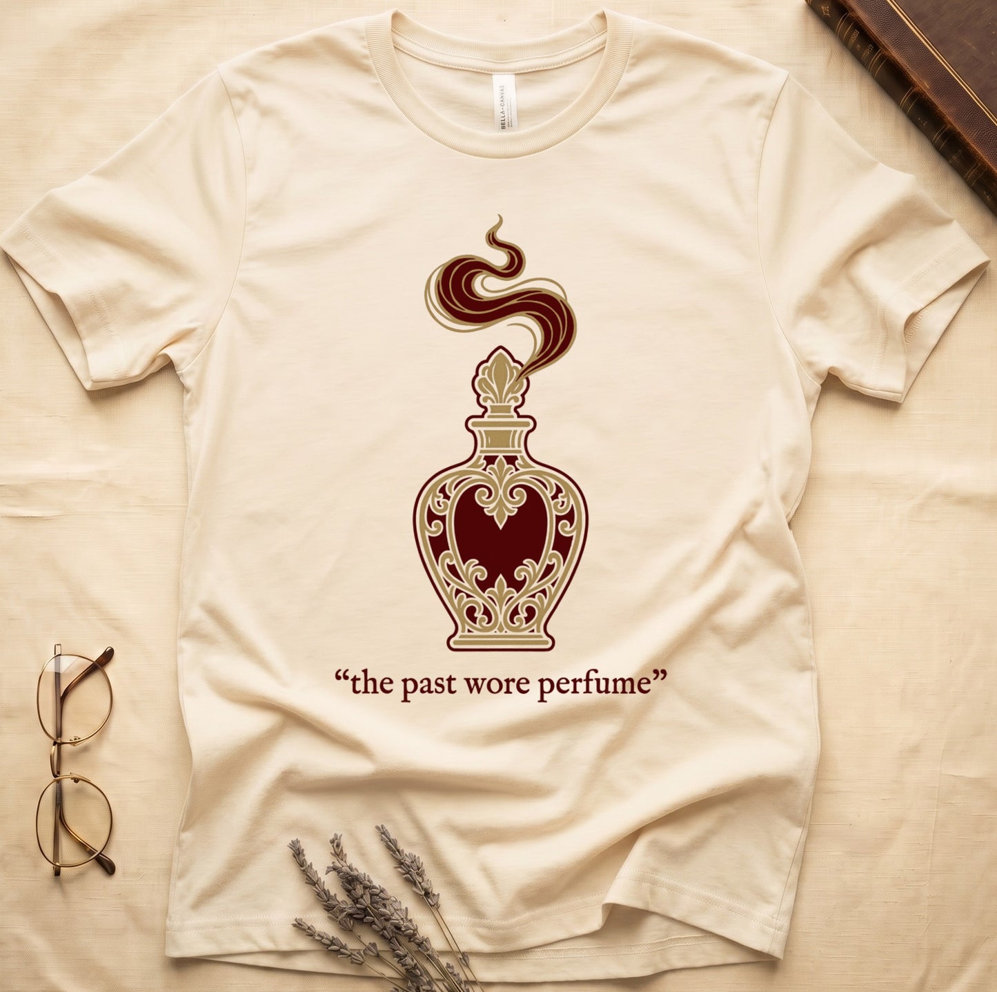 Natural tee with ornate blue and gold perfume bottle, rising scent swirl, "the past wore perfume" cream serif below.