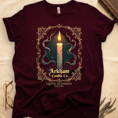 Maroon tee with lit candle, smoke, ornate gold filigree border, "Arkham Candle Co.," "Lighting the Darkness Since 1920."