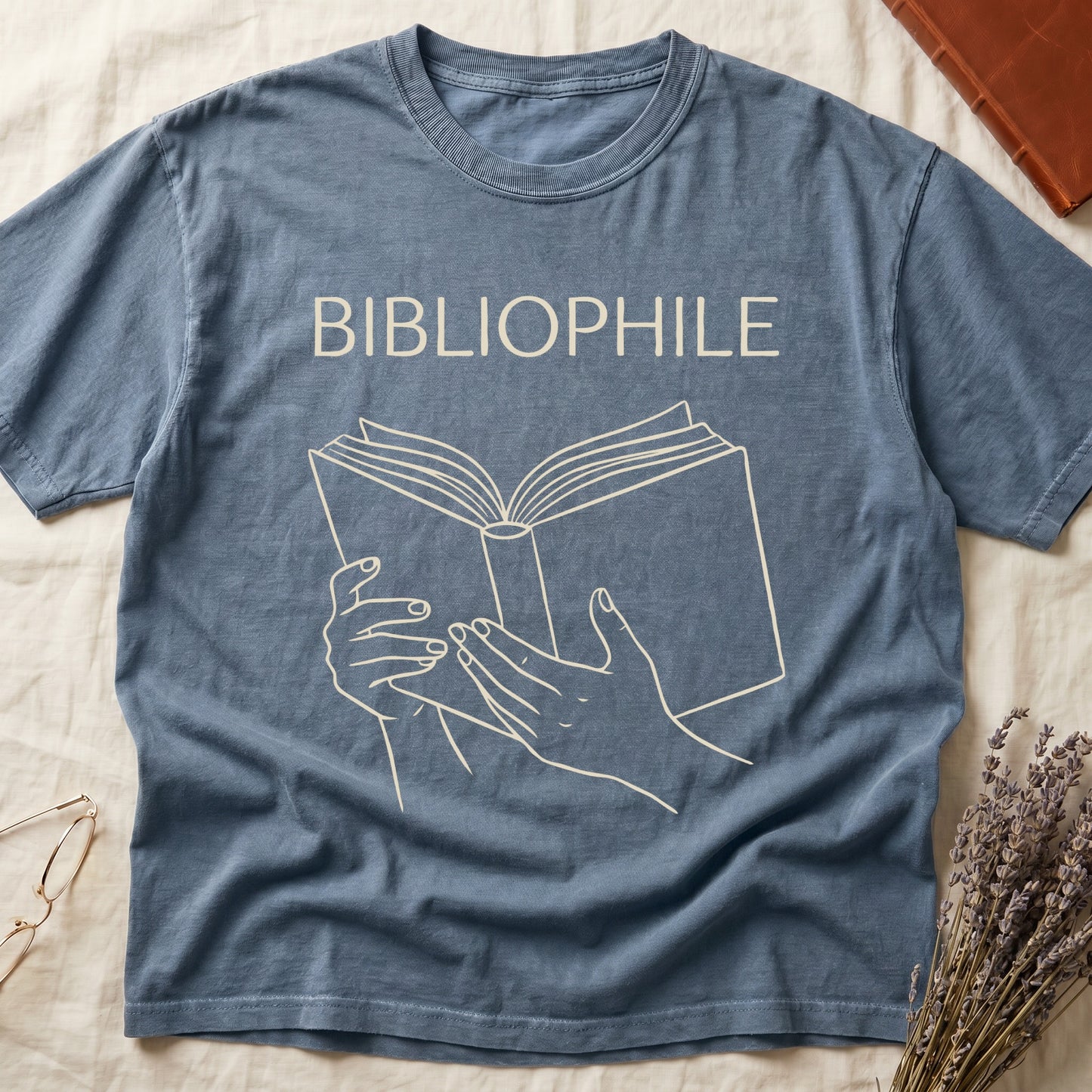 Blue Jean tee with two hands cradling an open book in cream and brown linework, "BIBLIOPHILE" in serif capitals above.
