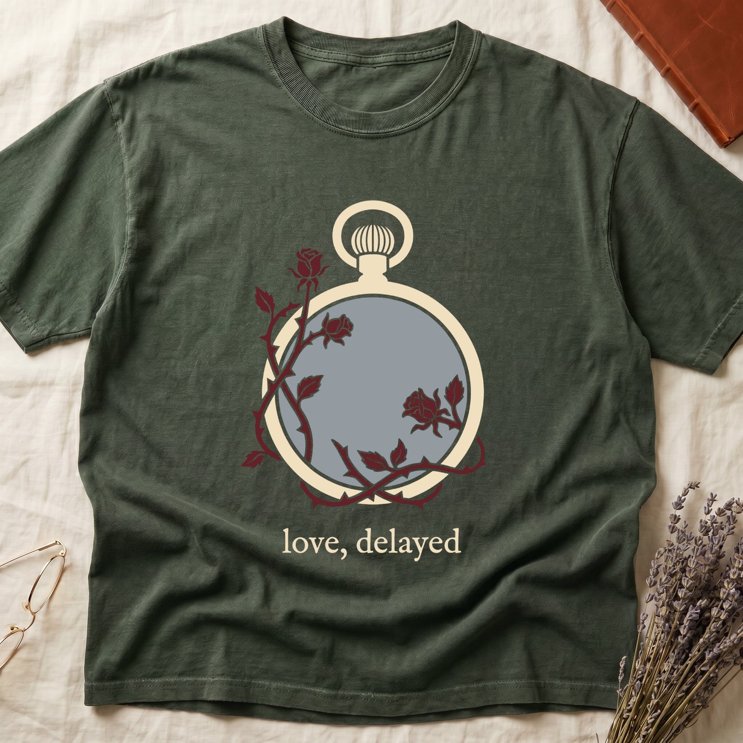 Green tee with vintage cream pocket watch and dark red roses entwined, "love, delayed" in cream serif below.